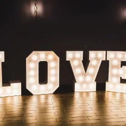 Duże, podświetlane litery tworzące słowo LOVE, ustawione na drewnianej podłodze w zaciemnionym pomieszczeniu z widocznymi kinkietami na ścianie w tle.
