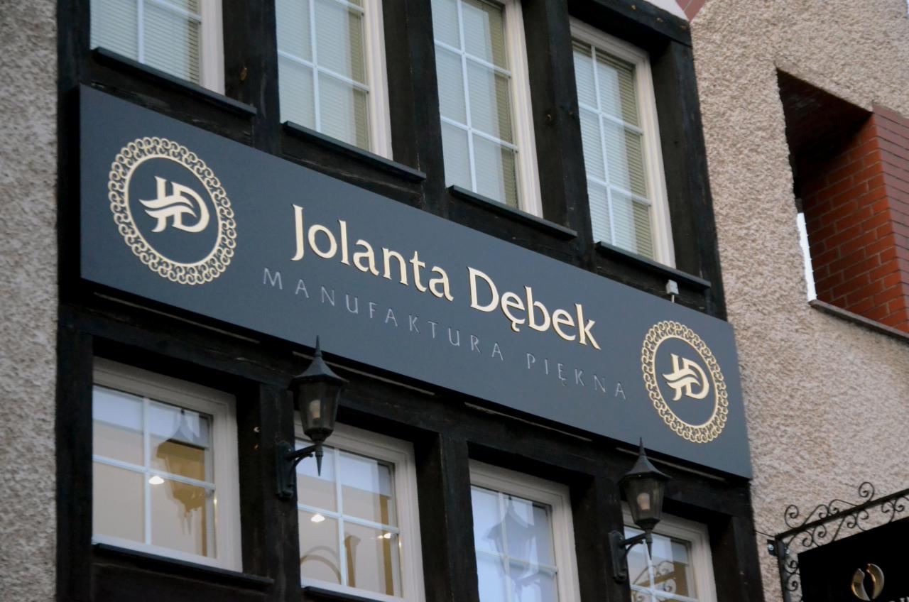 Szyld firmy Jolanta Dębek Manufaktura Piękna w kolorze grafitowym ze złotymi napisami i logo, zamontowany na elewacji budynku z oknami i czarnymi elementami dekoracyjnymi.