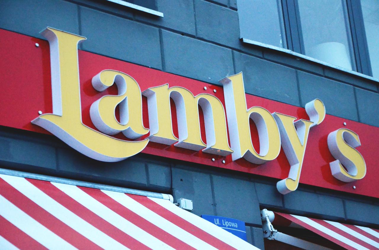 Zewnętrzny szyld z nazwą 'Lamby's' w kolorze żółto-białym na czerwonym tle, zamontowany na szarej ścianie budynku, z widocznym fragmentem markizy w biało-czerwone pasy i tabliczką z nazwą ulicy 'Ul...