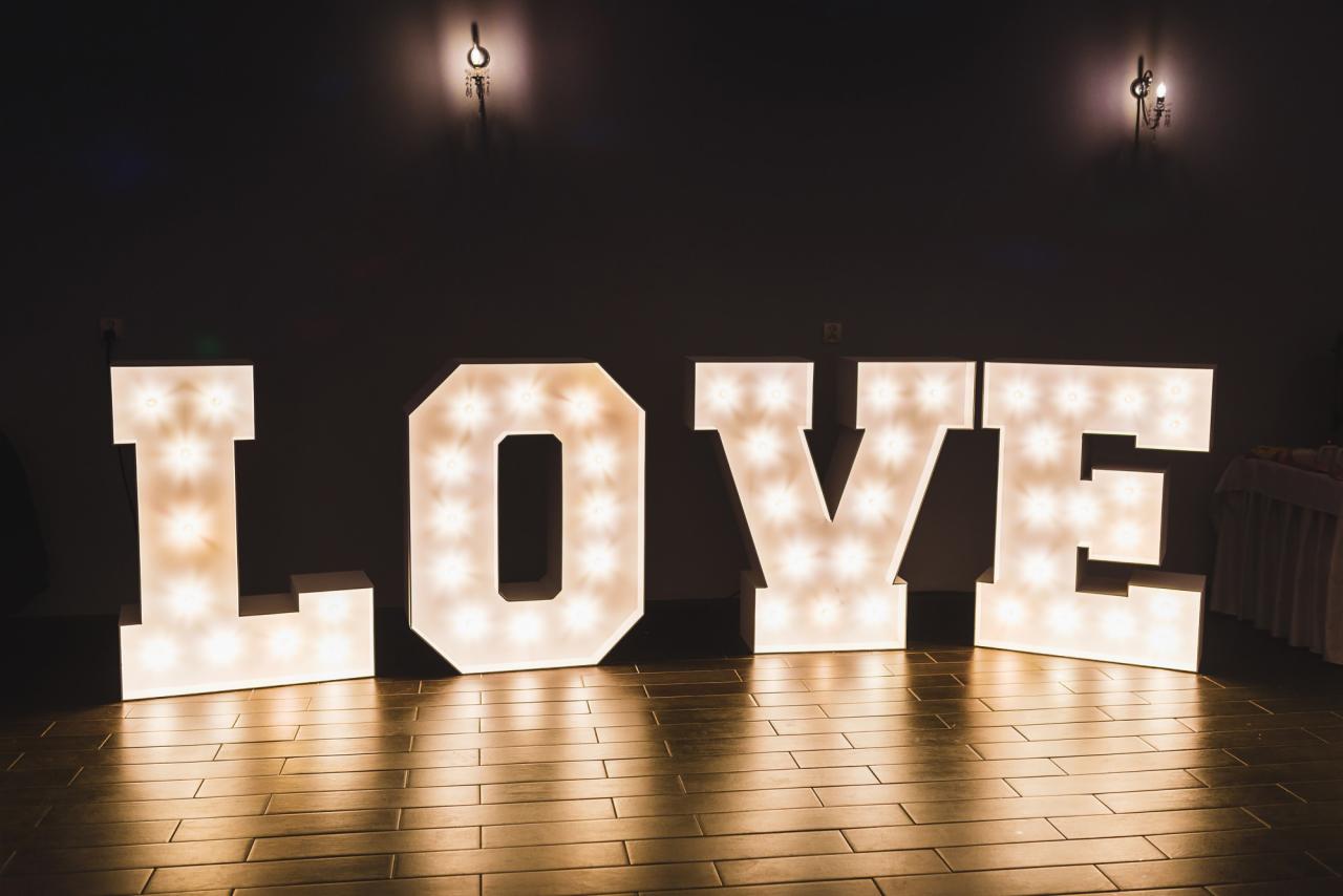 Duże, podświetlane litery tworzące słowo LOVE, ustawione na drewnianej podłodze w zaciemnionym pomieszczeniu z widocznymi kinkietami na ścianie w tle.