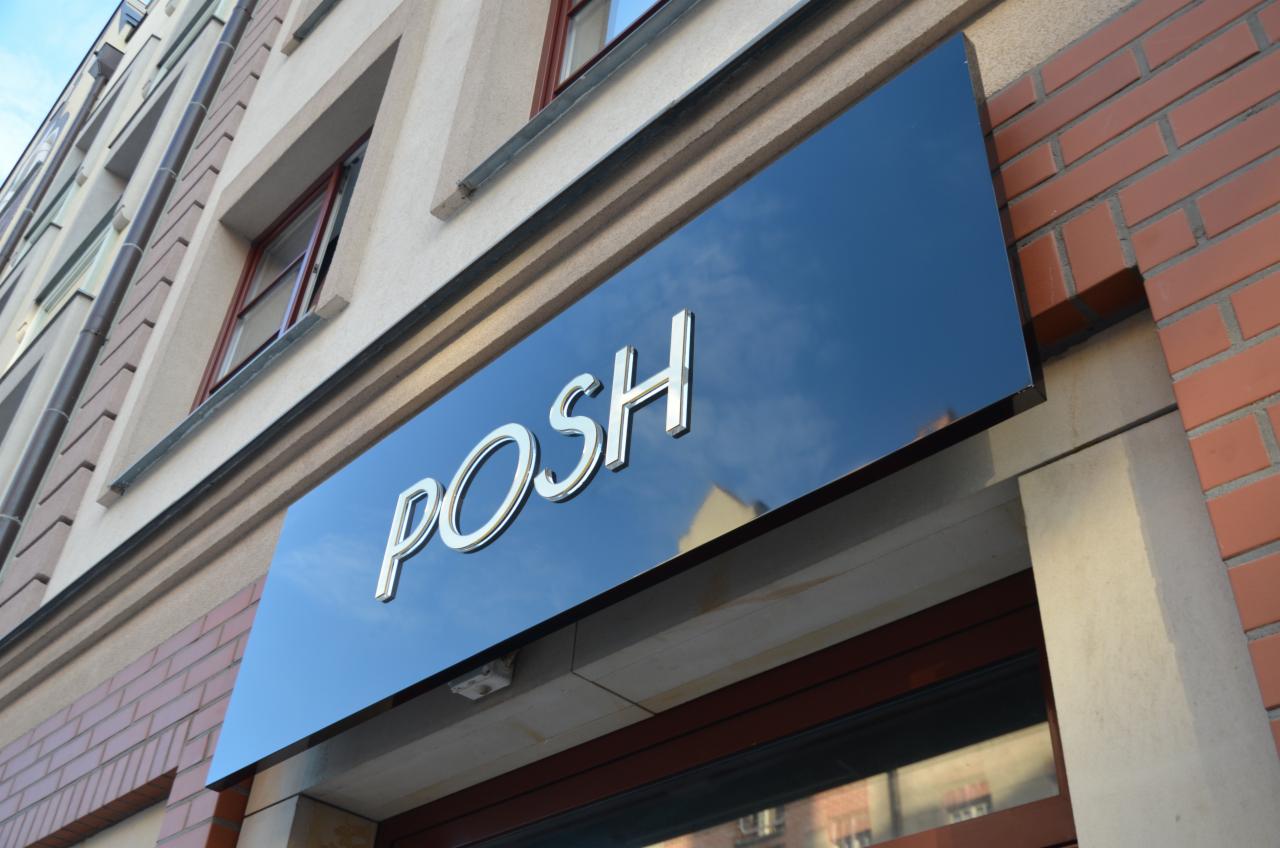 Błyszczący, czarny szyld z chromowanymi literami 'POSH' zamontowany nad wejściem do budynku z ceglaną elewacją.