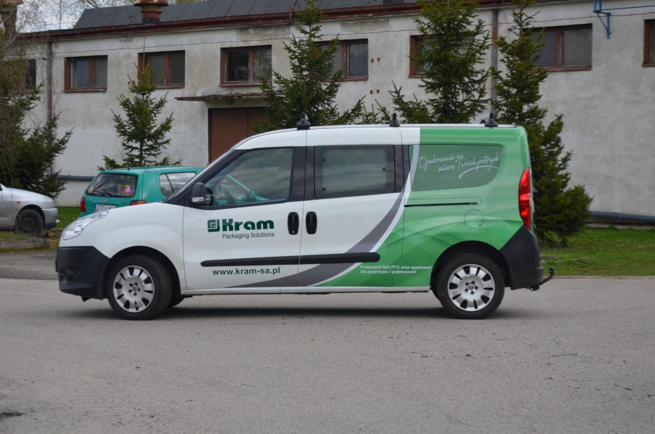 Biało-zielony samochód dostawczy z logo firmy produkującej folie PVC i opakowania dla przemysłu i gastronomii, zaparkowany na asfaltowej drodze przed budynkiem.