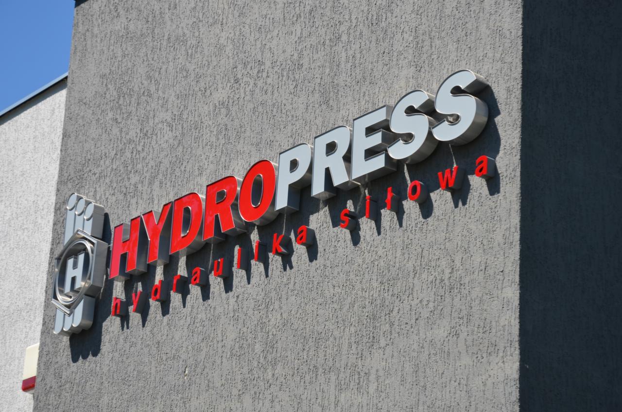 Zewnętrzny szyld firmy hydraulicznej z napisem 'HYDROPPRESS' w kolorze czerwono-białym, zamontowany na szarej, teksturowanej elewacji budynku w słoneczny dzień.