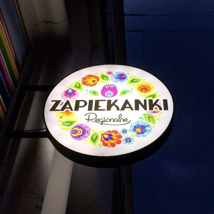 Podświetlany, okrągły szyld z napisem 'ZAPIEKANKI Regionalne' otoczony kolorowymi, folkowymi motywami kwiatowymi, zamontowany na zewnątrz budynku.