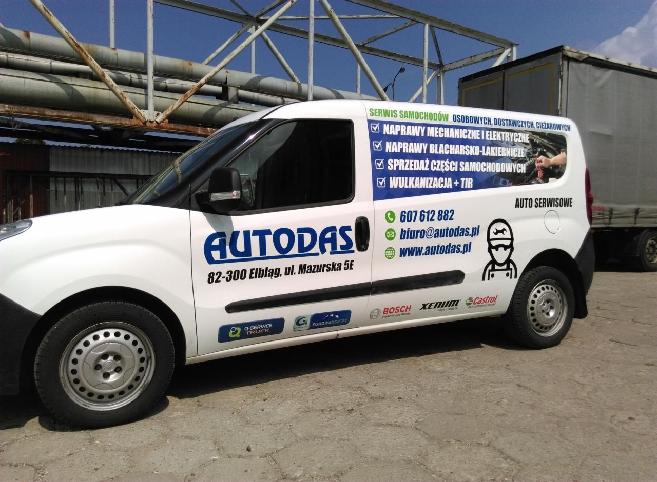 Biały samochód dostawczy z niebieskim logo firmy Autodas oferującej naprawy mechaniczne, blacharsko-lakiernicze, sprzedaż części samochodowych i wulkanizację, zaparkowany na zewnątrz w dzień.