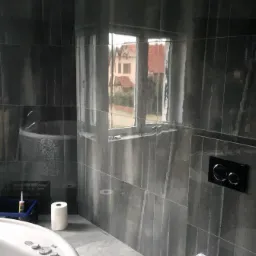 Wykończona łazienka z ciemnoszarymi, pionowo ułożonymi płytkami imitującymi kamień, z widoczną wanną z hydromasażem i toaletą, okno z widokiem na dom z czerwonym dachem.