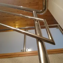 Spawacz poleruje metalową balustradę schodów z wypełnieniem ze szkła mlecznego, widoczny drewniany sufit i fragment schodów.