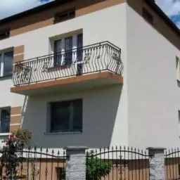 Dwupiętrowy dom z jasną elewacją i balkonem z ozdobną, kutą balustradą. Widoczne brązowe obramowania okien i górnej części elewacji. W tle błękitne niebo z chmurami.