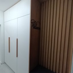 WoodMood Design Adam Pańkow - Biała szafa wnękowa z długimi, pionowymi, drewnianymi uchwytami, sąsiadująca z drewnianym panelem ściennym z siedziskiem i dekoracją 'Welcome' na ścianie.