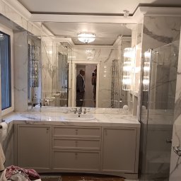 WoodMood Design Adam Pańkow - Łazienka w trakcie wykańczania z szarą szafką pod umywalkę z trzema szufladami i lustrem składającym się z wielu tafli, widoczna osoba w odbiciu.