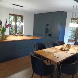WoodMood Design Adam Pańkow - Nowoczesna kuchnia z wyspą i drewnianym blatem, szare szafki, drewniany stół z welurowymi krzesłami, ozdobne lampy wiszące i kwiaty w wazonie.