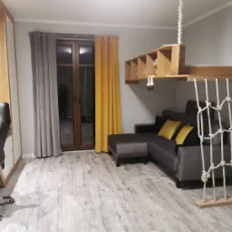 Pokój z szarą sofą narożną, drewnianą półką podwieszoną pod sufitem na linach oraz drabinką sznurową, na podłodze panele imitujące bielone drewno.