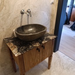 WoodMood Design Adam Pańkow - Kamienna umywalka w kształcie misy na drewnianej szafce z blatem z granitu, chromowana bateria ścienna, beżowe płytki na ścianie i podłodze, widok na otwarte drzwi do pomieszczenia.
