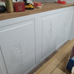 WoodMood Design Adam Pańkow - Białe fronty szafek z frezowanymi motywami kwiatowymi lilii, zamontowane pod blatem z widocznymi narzędziami i akcesoriami montażowymi, w trakcie prac wykończeniowych.