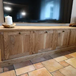 Drewniana szafka RTV z dekoracyjnymi frezami i okrągłymi uchwytami, na blacie świeca i drewniana ozdoba, w tle telewizor i głośnik.