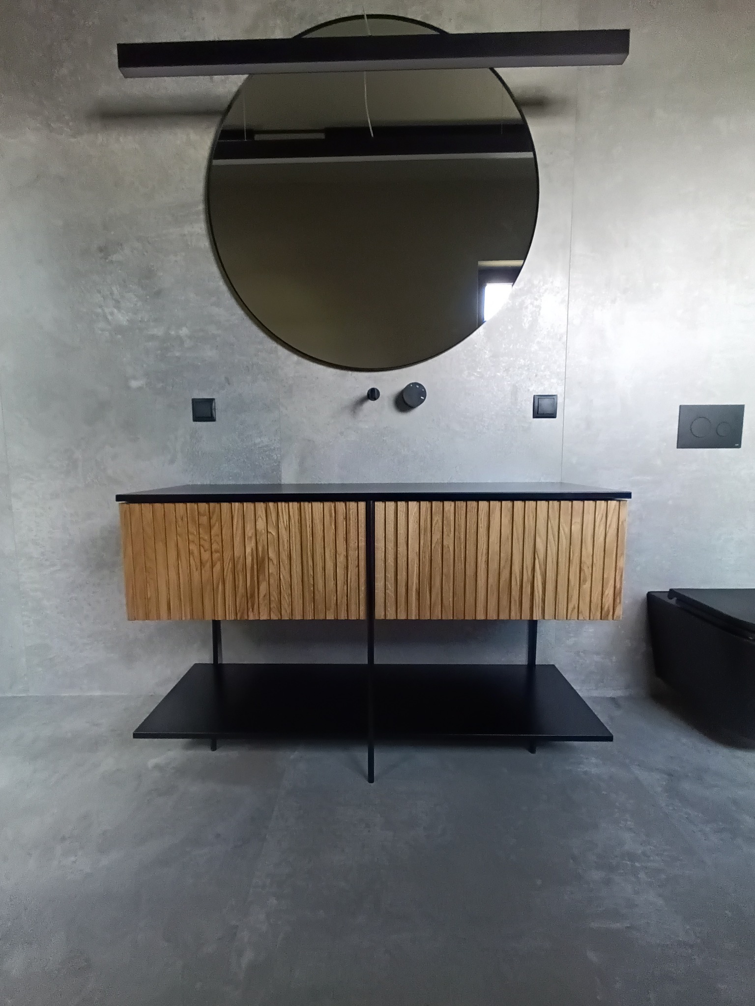 Nowoczesna łazienka z drewnianą szafką pod umywalkę o ryflowanych frontach, okrągłym lustrem i betonową ścianą w tle.