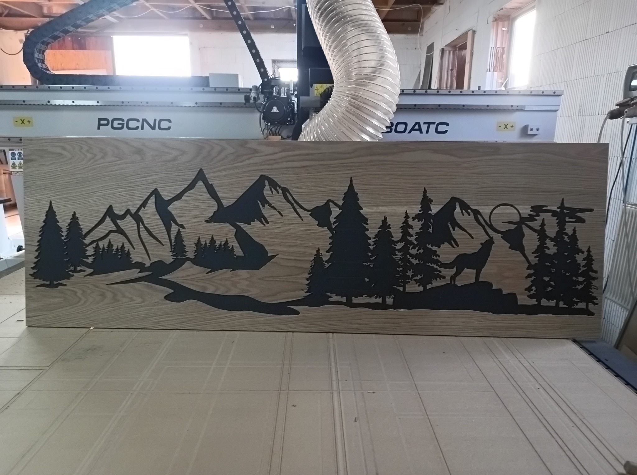 Płyta z jasnego drewna z wyciętym wzorem górskiego krajobrazu z wilkiem, leżąca na stole frezarki CNC PGCNC 30ATC.
