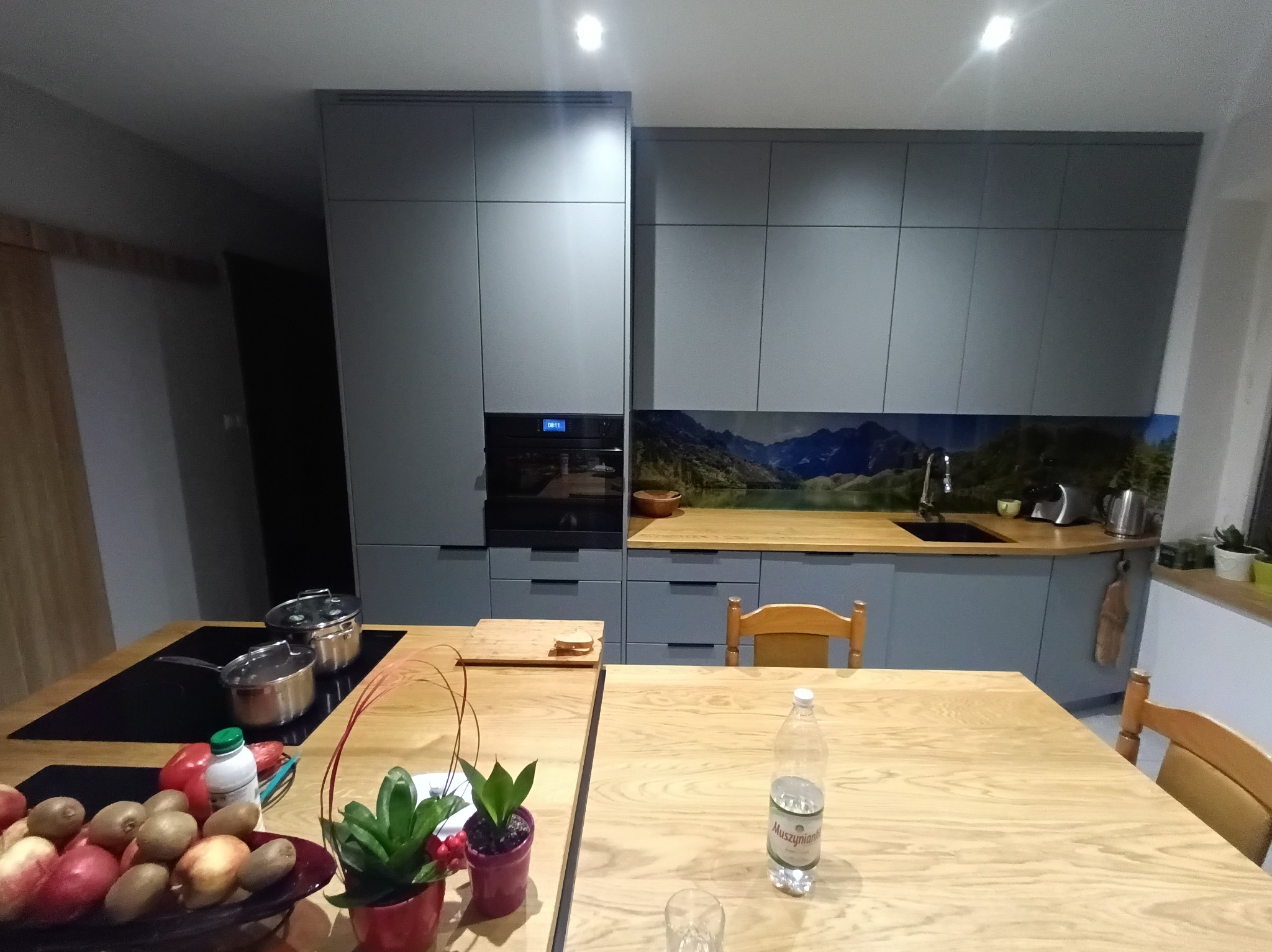 Nowoczesna kuchnia z szarymi szafkami na wymiar, drewnianym blatem i panelem ściennym z motywem górskim, na pierwszym planie stół z owocami, garnkami i butelką wody.