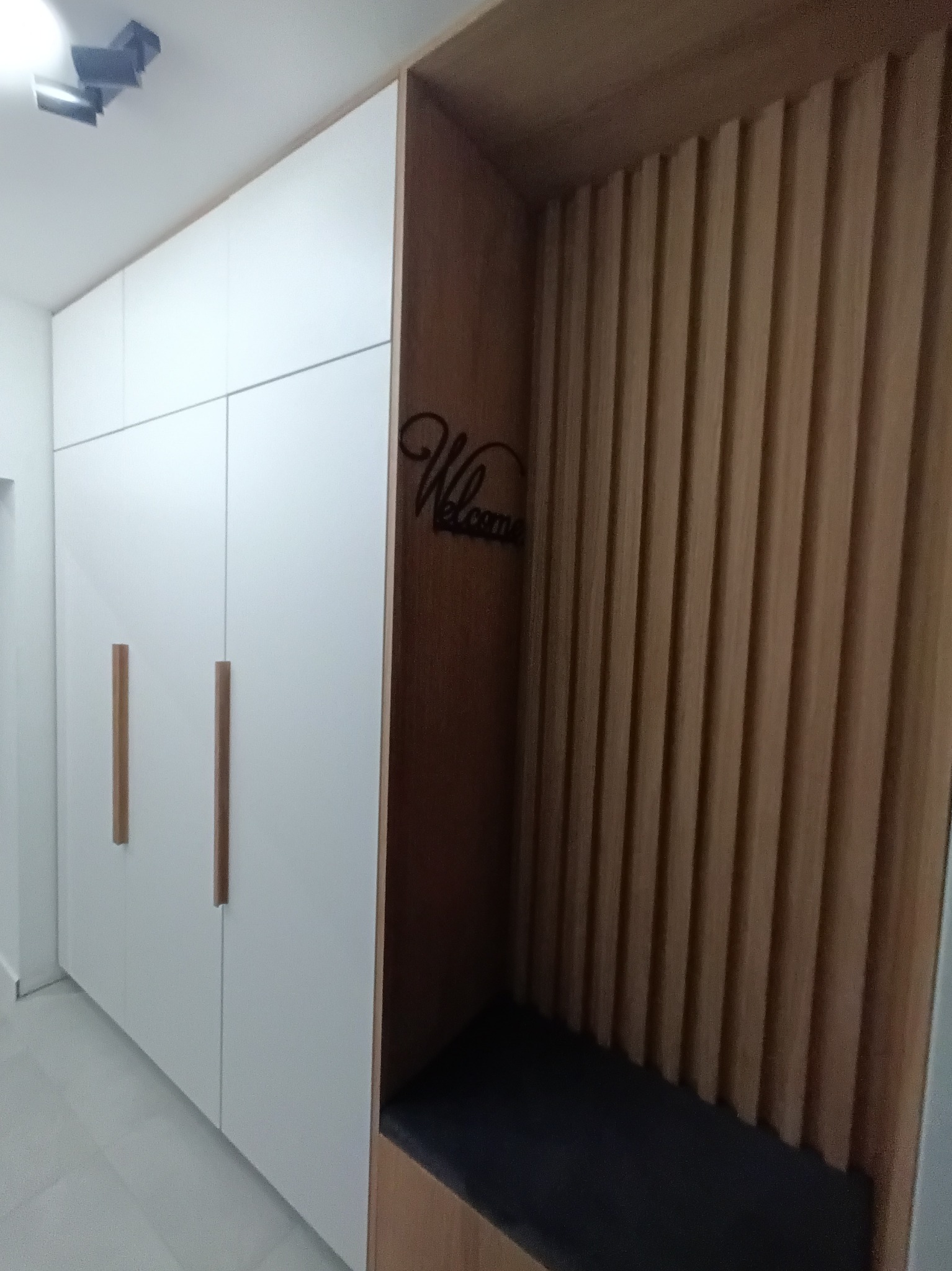 Biała szafa wnękowa z długimi, pionowymi, drewnianymi uchwytami, sąsiadująca z drewnianym panelem ściennym z siedziskiem i dekoracją 'Welcome' na ścianie.