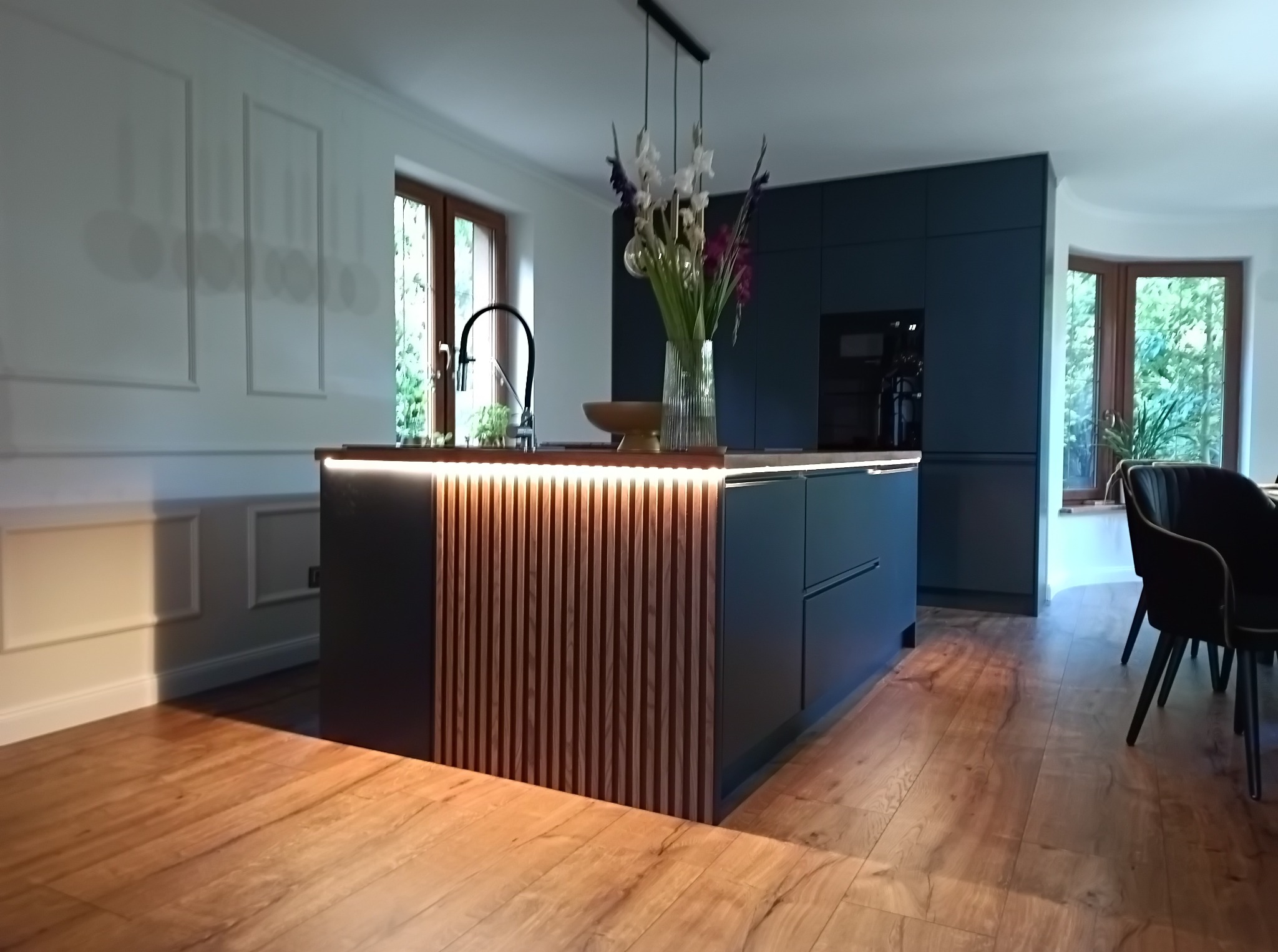 Nowoczesna wyspa kuchenna z ciemnymi szafkami, drewnianą okładziną z pionowych listew podświetlonych od góry, drewnianym blatem, kwiatami w wazonie i drewnianą podłogą w jasnym wnętrzu.