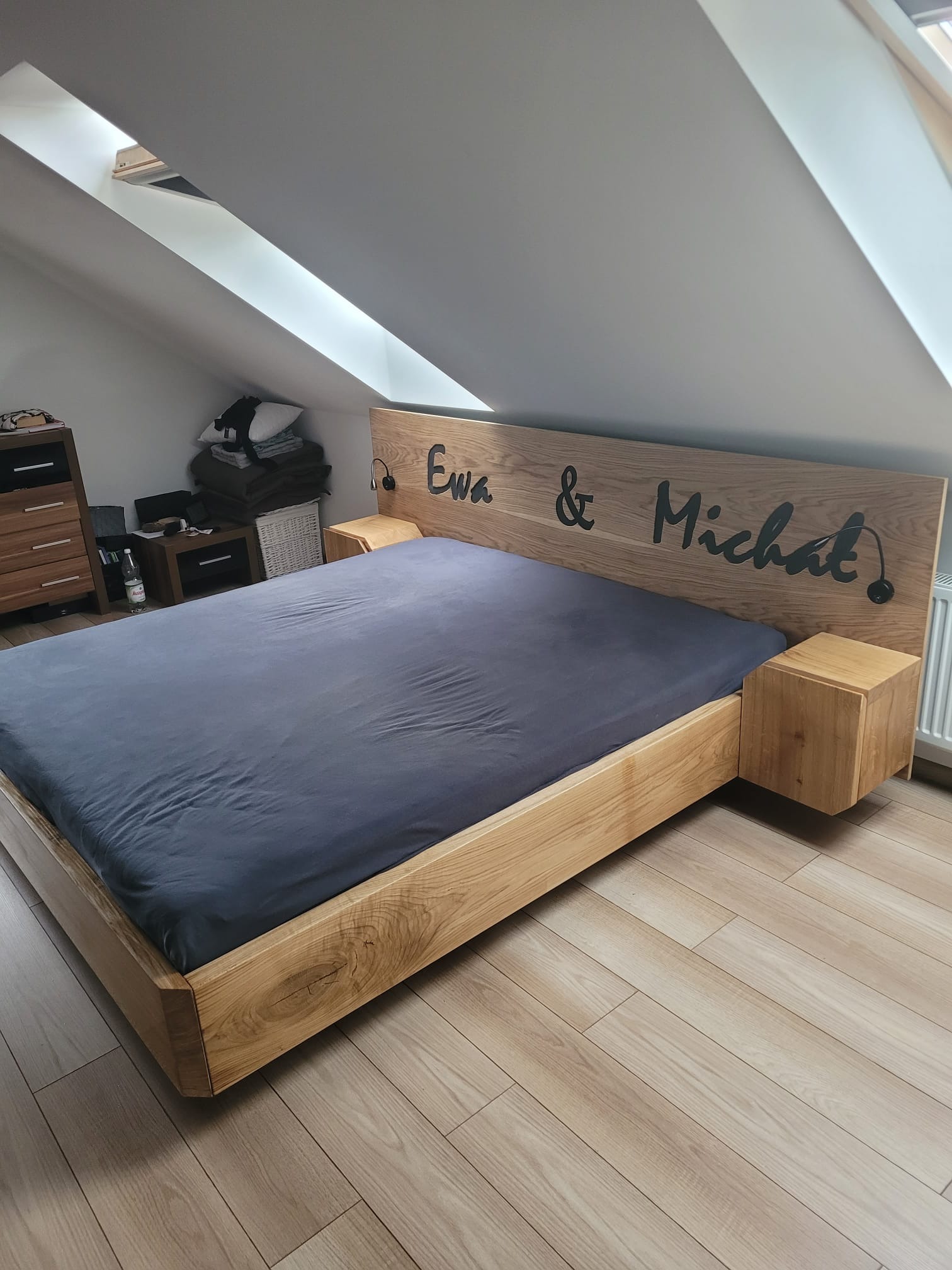 Drewniane łóżko małżeńskie z zagłówkiem z napisem 'Ewa & Michał' w pokoju na poddaszu z oknami dachowymi i drewnianą podłogą.
