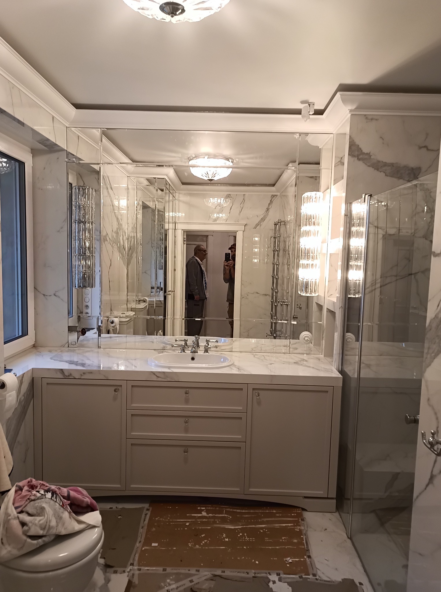 Łazienka w trakcie wykańczania z szarą szafką pod umywalkę z trzema szufladami i lustrem składającym się z wielu tafli, widoczna osoba w odbiciu.