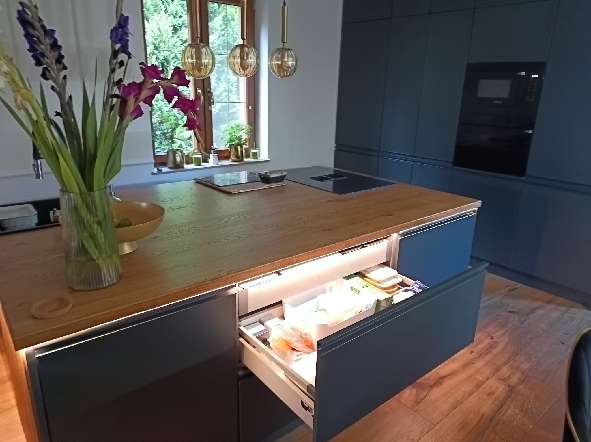 Nowoczesna kuchnia z szarymi szafkami i drewnianym blatem, otwartą szufladą z oświetleniem LED, w której widać zapasy żywności, oraz bukietem kwiatów w szklanym wazonie na blacie.