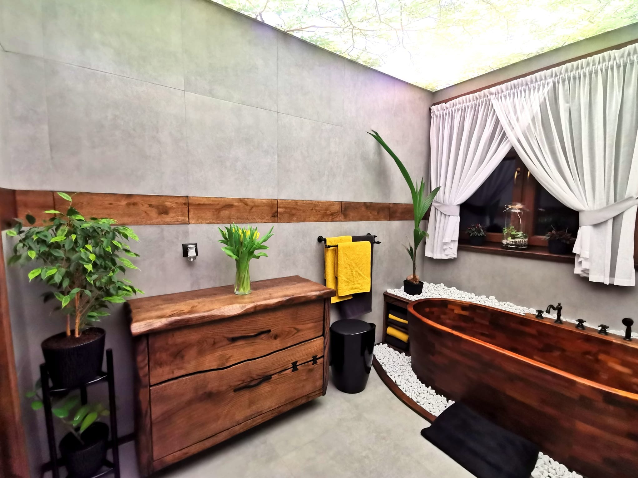 Rustykalna łazienka z drewnianą wanną i komodą z litego drewna, żółte tulipany w wazonie, sufit z motywem liści, szare płytki i białe zasłony.