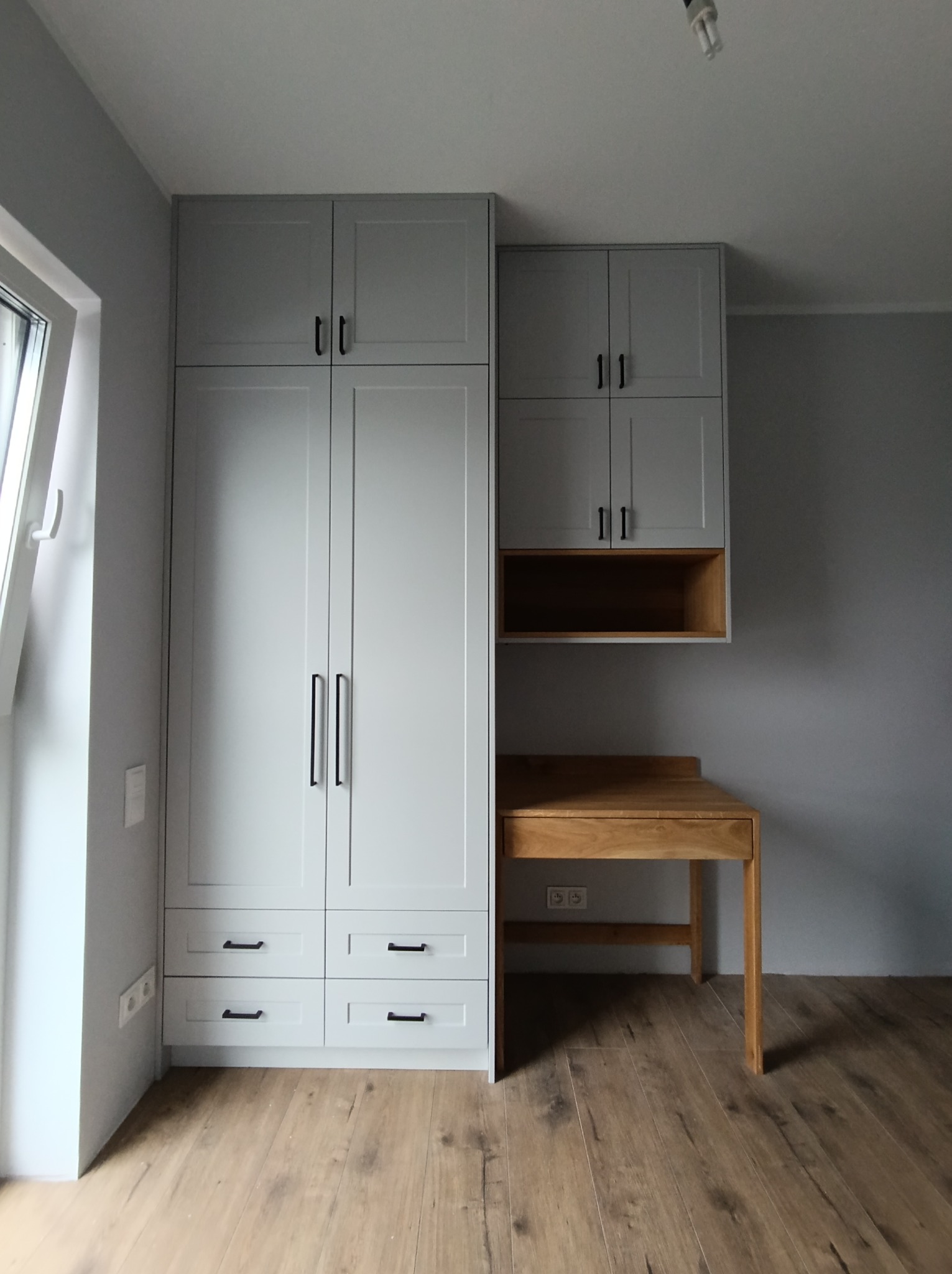 Szara zabudowa meblowa z szafą, szafkami wiszącymi i biurkiem z jasnego drewna na tle szarej ściany i drewnianej podłogi.