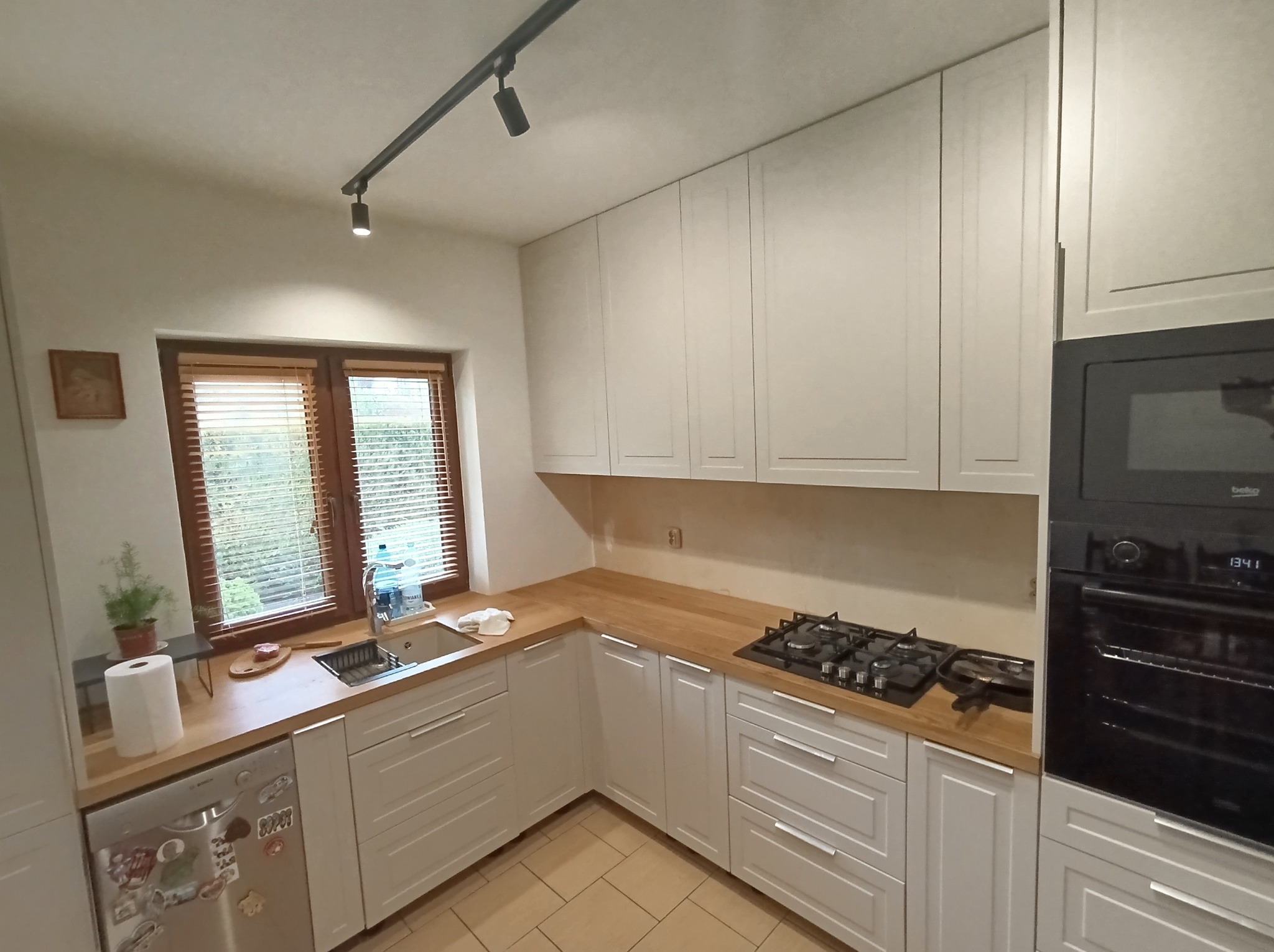 Jasna kuchnia z białymi szafkami, drewnianym blatem i oknem z żaluzjami, widok na zlew, płytę gazową i piekarnik.