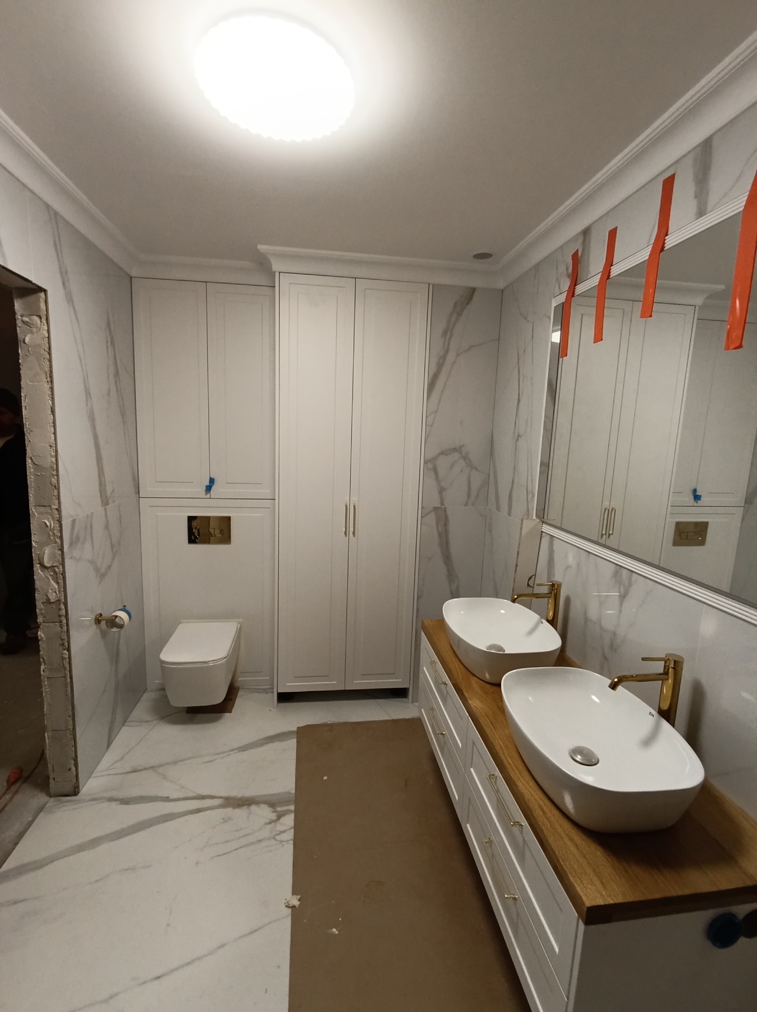 Nowoczesna łazienka w trakcie wykończenia z marmurowymi płytkami, podwójną umywalką nablatową na drewnianym blacie, białą zabudową meblową od podłogi do sufitu i wiszącą toaletą z złotym przyciskiem.