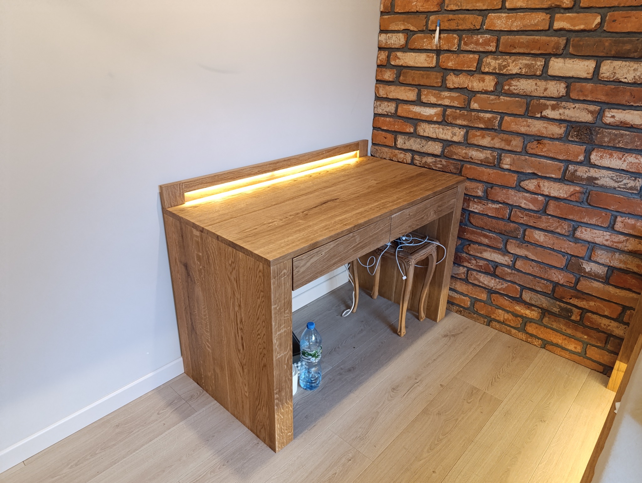 Biurko z oświetleniem LED wbudowanym w blat, z krzesłem w stylu retro, ustawione na tle ściany z cegły i białej ściany. Pod biurkiem widoczna butelka wody.