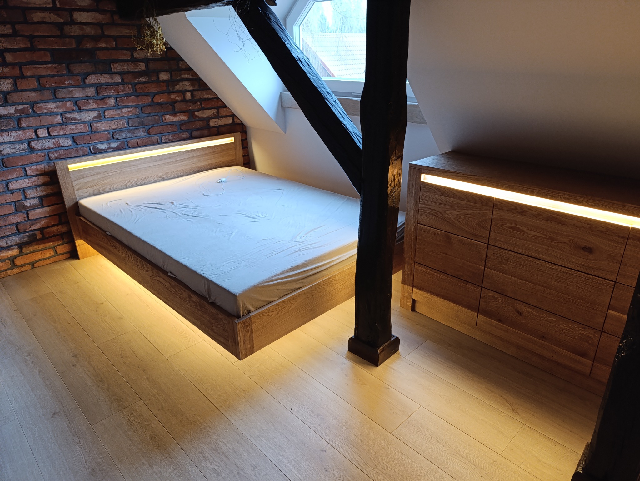Sypialnia na poddaszu z drewnianym łóżkiem podświetlanym od spodu i komodą z oświetleniem LED, widoczna ceglana ściana i element konstrukcyjny z ciemnego drewna.