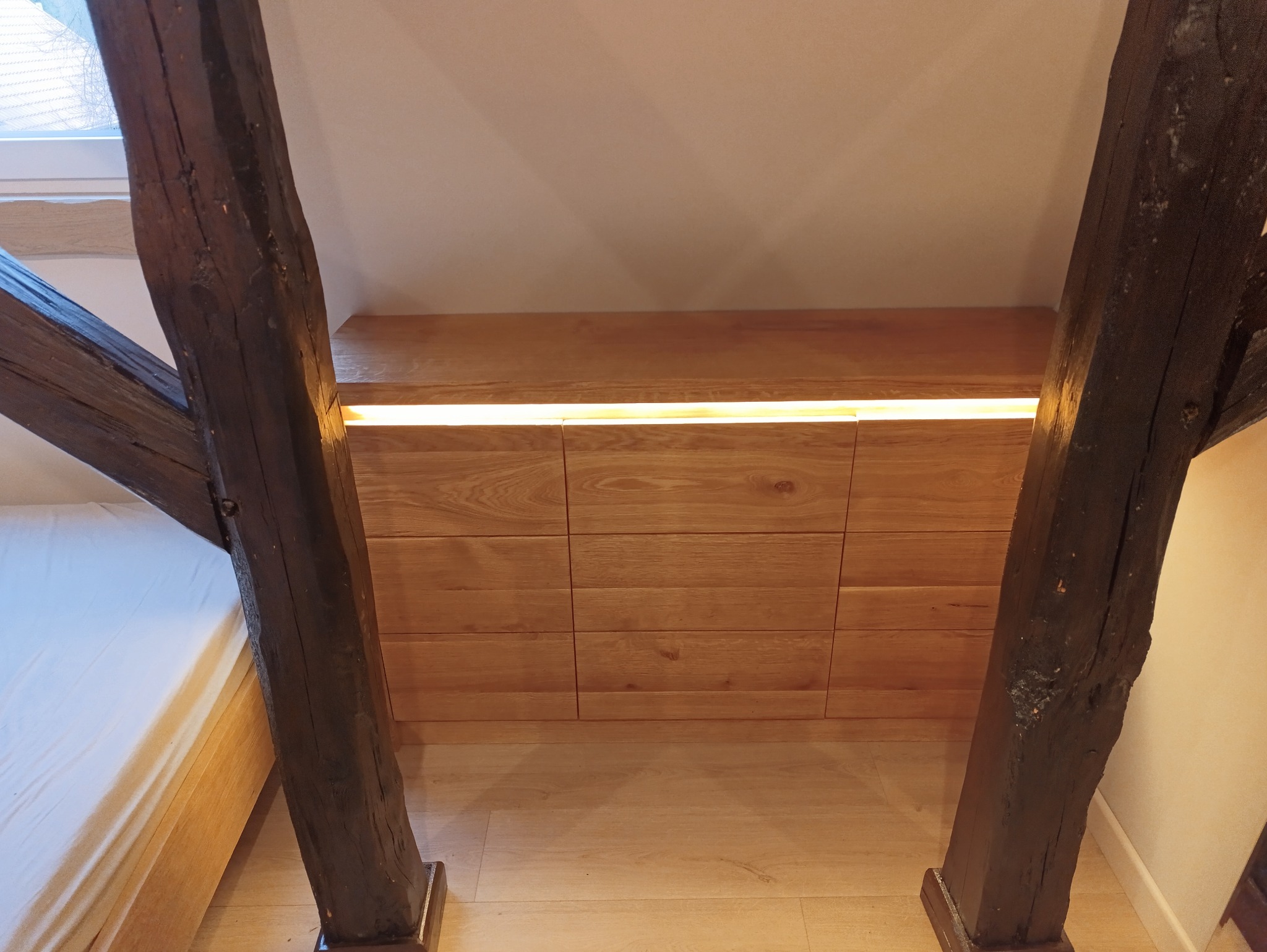 Szafa wkomponowana w skos dachu z drewnianymi belkami konstrukcyjnymi, oświetlona listwą LED, widok z dołu, sypialnia na poddaszu.