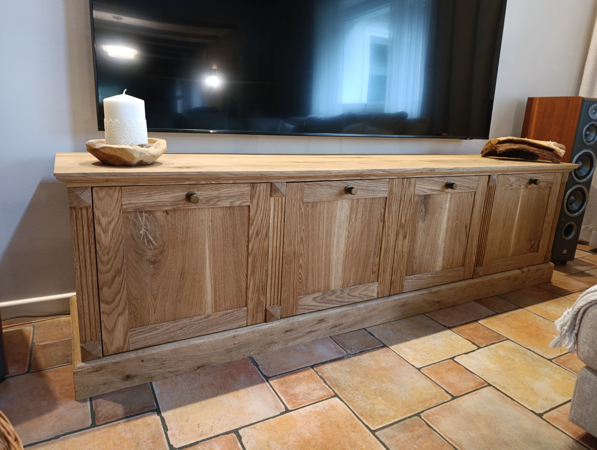 Drewniana szafka RTV z dekoracyjnymi frezami i okrągłymi uchwytami, na blacie świeca i drewniana ozdoba, w tle telewizor i głośnik.