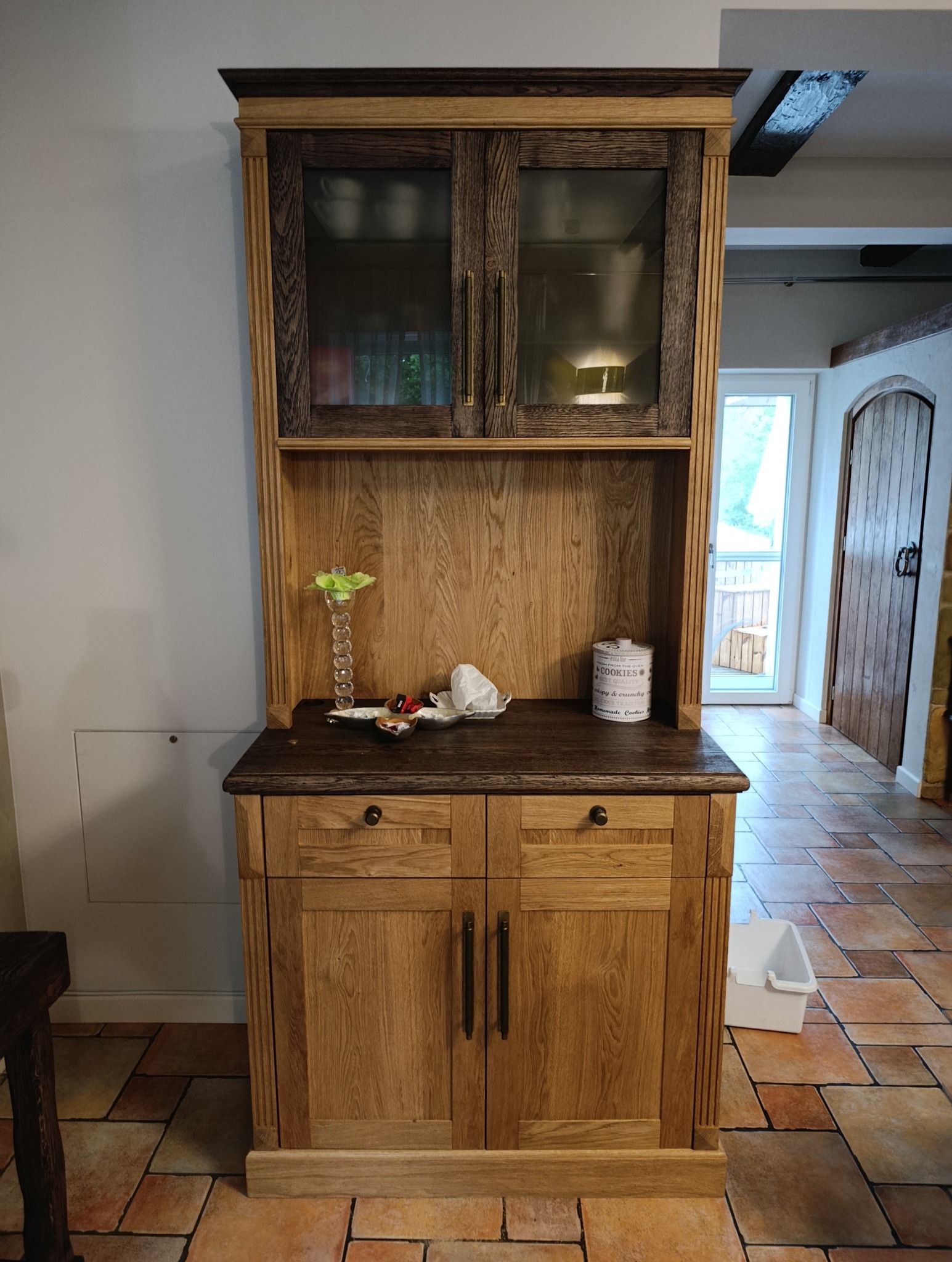 Dwukolorowy kredens z drewna dębowego, z przeszkloną górną częścią i ciemnym blatem, stoi na terakotowej podłodze w jasnym wnętrzu.