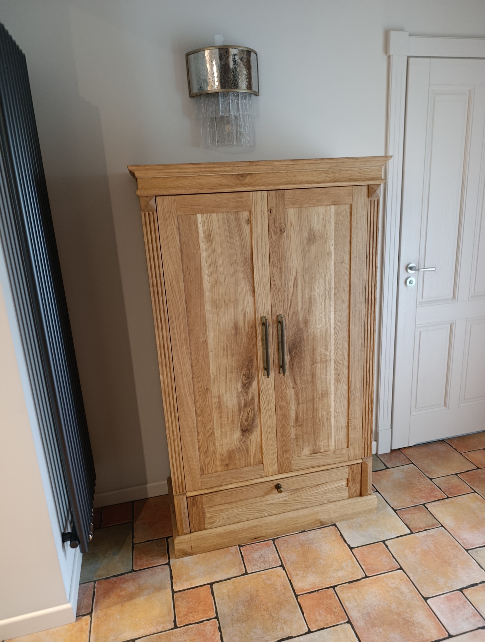 Drewniana szafa dwudrzwiowa z szufladą na tle terakoty, obok grzejnika i drzwi. Szafa z jasnego drewna z ciemnymi uchwytami, dekoracyjne frezy na bokach.