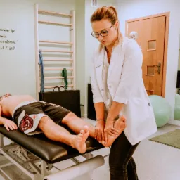 Kobieta w białym fartuchu uciska stopę pacjenta leżącego na czarnym stole do masażu w gabinecie z drabinką gimnastyczną i cytatem na ścianie.