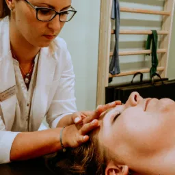 Kobieta w białym fartuchu, z identyfikatorem fizjoterapeuty, delikatnie masuje czoło pacjentki leżącej na leżance w gabinecie z drabinkami gimnastycznymi w tle.