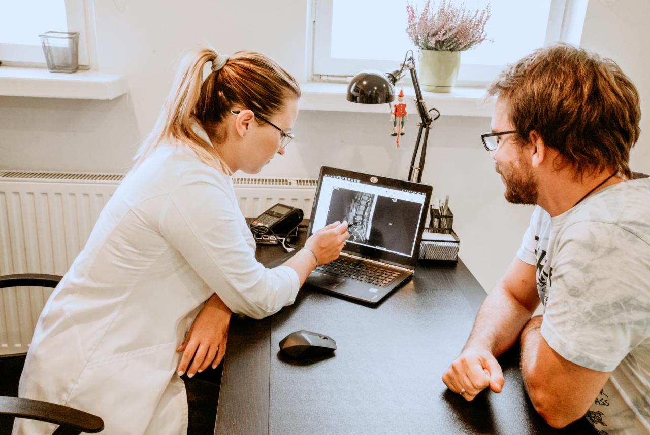 Lekarz w białym fartuchu omawia z pacjentem zdjęcie RTG kręgosłupa na laptopie, stojącym na biurku w gabinecie lekarskim.