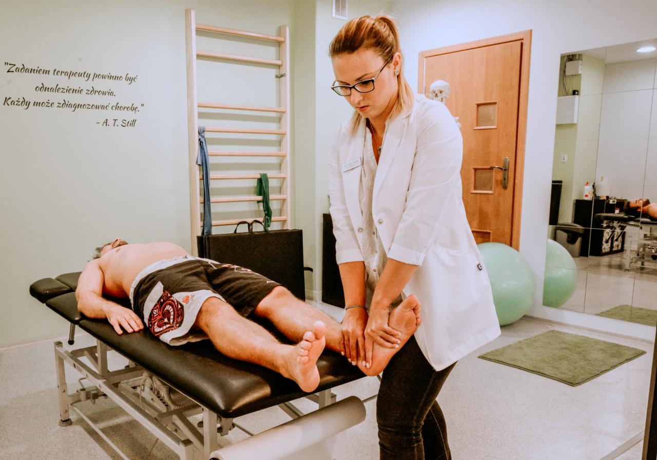 Kobieta w białym fartuchu uciska stopę pacjenta leżącego na czarnym stole do masażu w gabinecie z drabinką gimnastyczną i cytatem na ścianie.