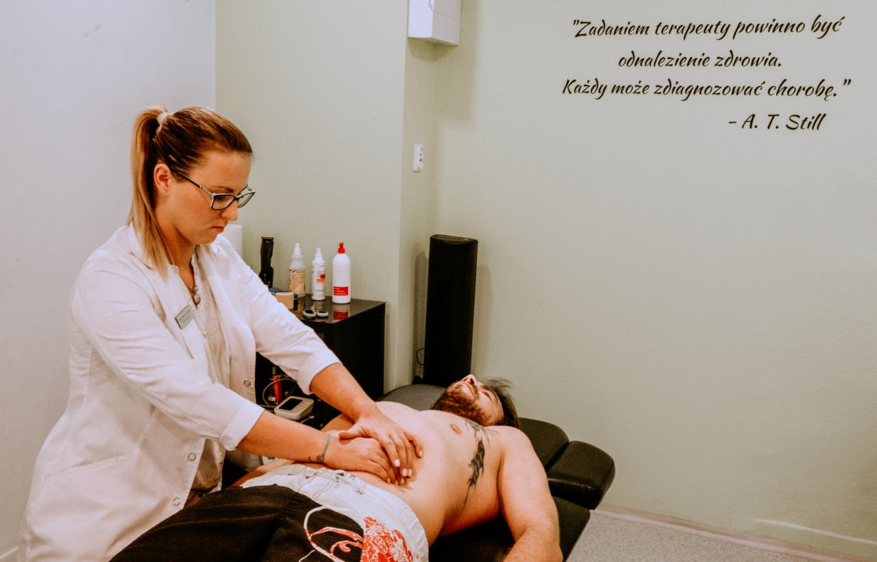 Kobieta w białym fartuchu uciska brzuch mężczyzny leżącego na czarnym stole do masażu; na ścianie widoczny cytat A.T. Stilla o terapii i zdrowiu.