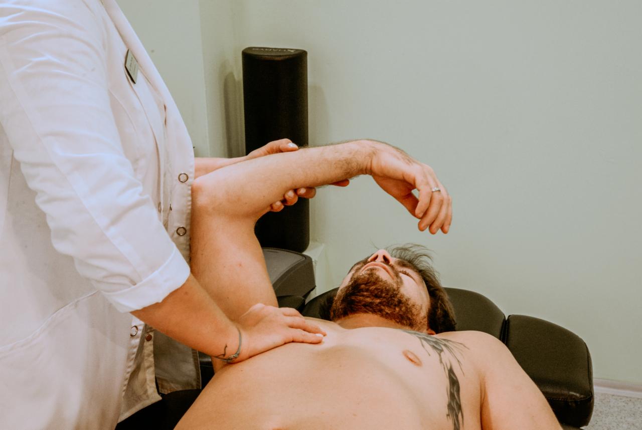 Pacjent leżący na kozetce podczas badania zakresu ruchu ramienia przez rehabilitanta w białym fartuchu.