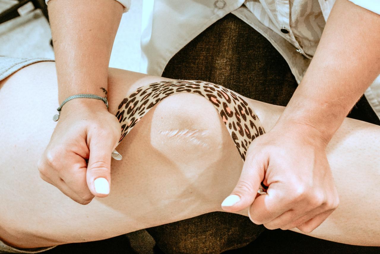 Nakładanie elastycznego plastra o wzorze panterki na kolano z widoczną blizną, jasne paznokcie, bransoletka na nadgarstku.
