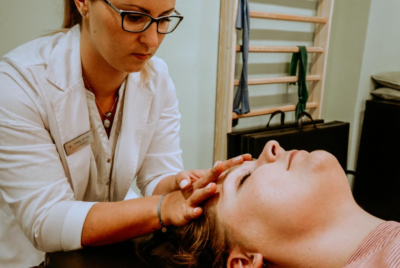 Kobieta w białym fartuchu, z identyfikatorem fizjoterapeuty, delikatnie masuje czoło pacjentki leżącej na leżance w gabinecie z drabinkami gimnastycznymi w tle.