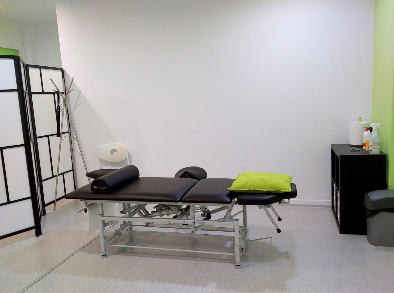 Pusty gabinet z profesjonalnym stołem do masażu z czarnym obiciem i jasnozieloną poduszką, parawanem pokojowym, krzesłem z wyciętym oparciem i czarną szafką z akcesoriami medycznymi.