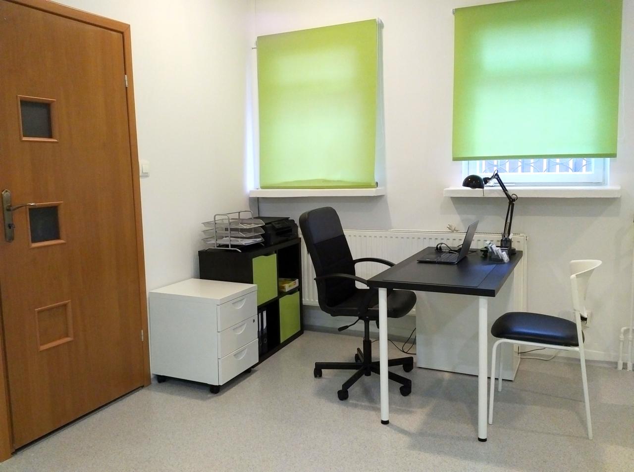 Gabinet rehabilitacyjny z biurkiem, krzesłami, szafkami i zielonymi roletami w oknach.