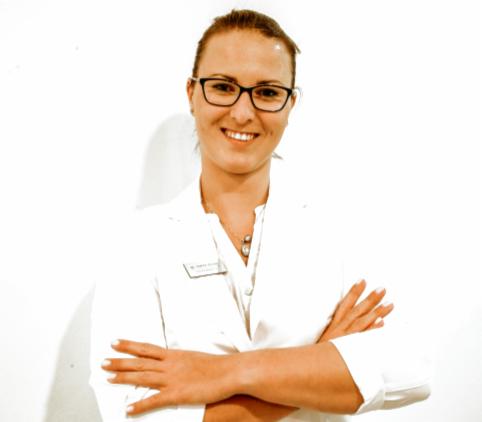 Uśmiechnięta kobieta w okularach i białym fartuchu medycznym, z założonymi rękami, stojąca na tle białej ściany.