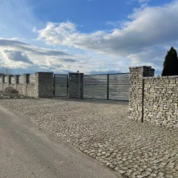Podjazd z kostki granitowej ułożonej w wachlarz, ogrodzenie z kamiennymi słupkami i metalowymi przęsłami, kamień z numerem 40b po lewej stronie.