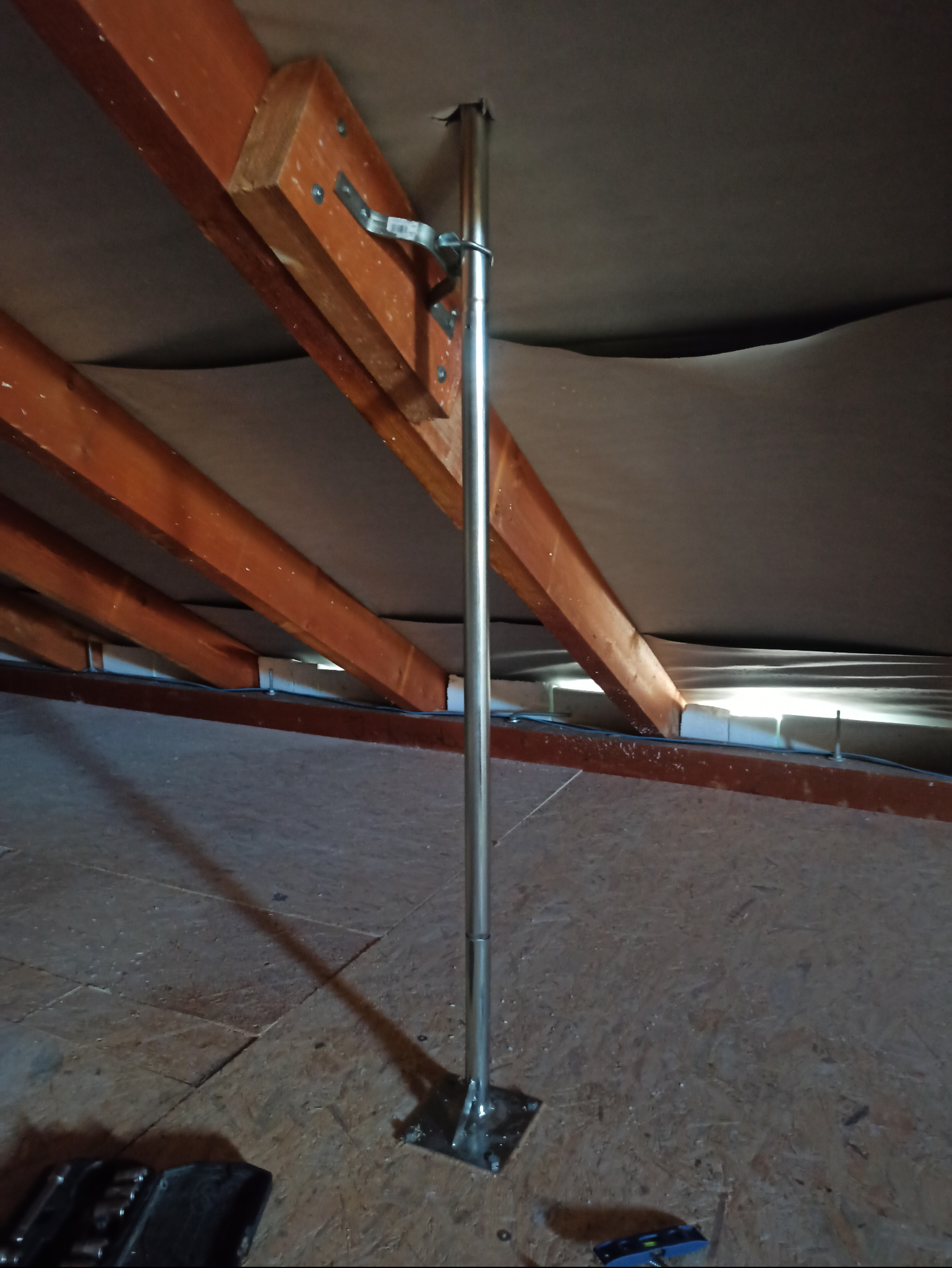 Metalowa rura antenowa zamocowana do drewnianej konstrukcji dachu na poddaszu, widoczne narzędzia w tle.